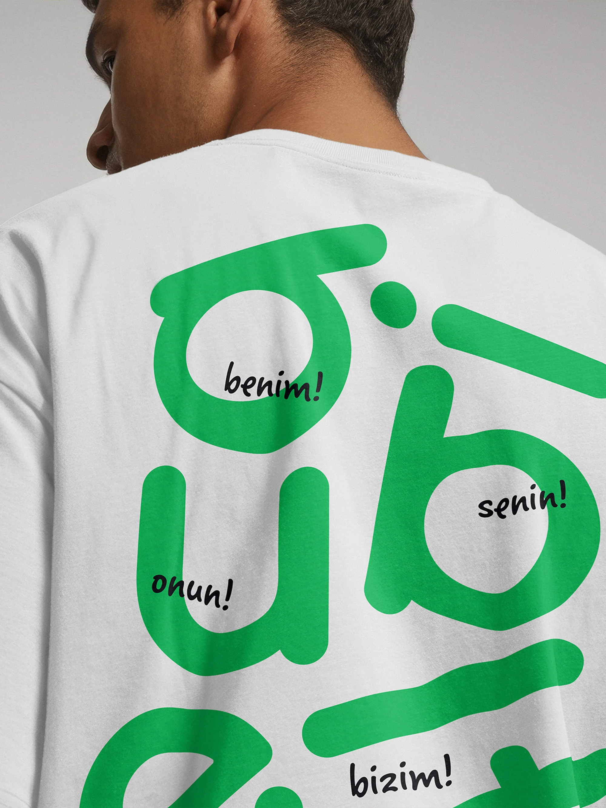Person facing away wearing a white t-shirt with green graphic text in Turkish, isolated on a white background in an indoor setting.