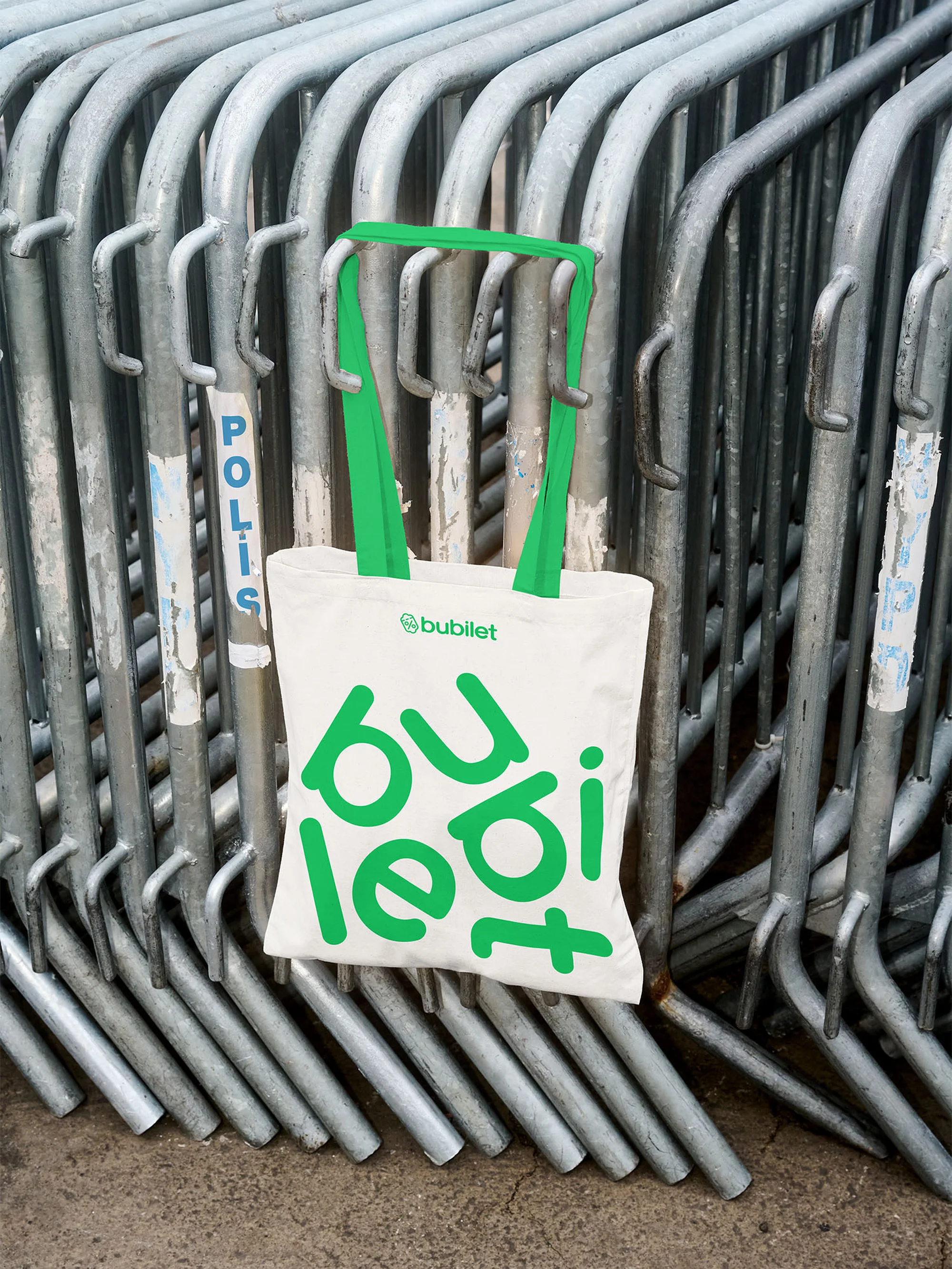 Tote bag with green handles and 'bubilet' logo hanging on metal crowd control barriers in an outdoor urban environment.