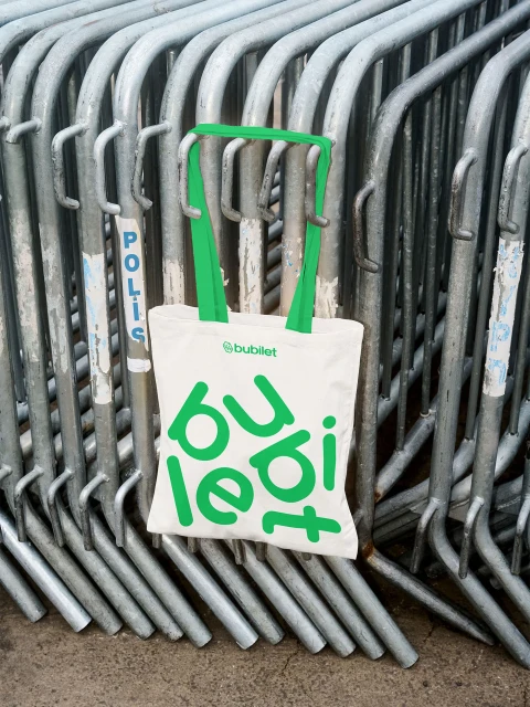 Tote bag with green handles and 'bubilet' logo hanging on metal crowd control barriers in an outdoor urban environment.