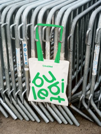 Tote bag with green handles and 'bubilet' logo hanging on metal crowd control barriers in an outdoor urban environment.
