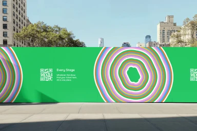 Green construction hoarding with concentric colorful rings and QR code outdoors under a clear sky.