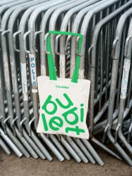 White tote bag with green handles and Bubilet branding hanging on stacked metal crowd control barriers.