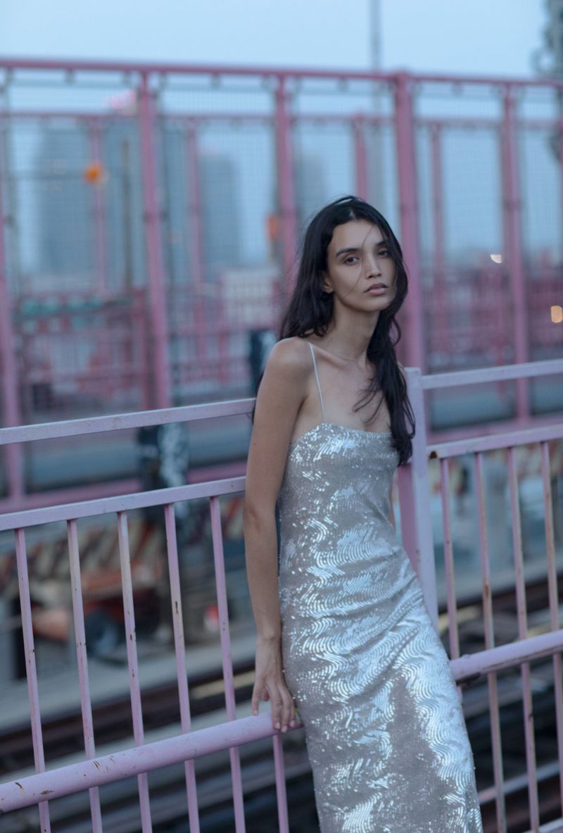 Elegant adult female in a sequined slip dress stands on a pink bridge with a blurred cityscape in the background in an urban setting.