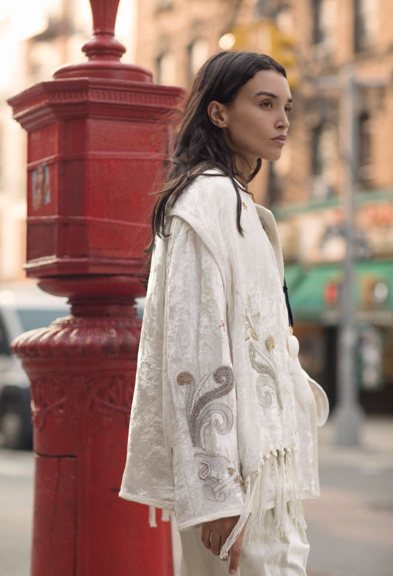 Woman wearing a patterned white jacket on a city street, standing next to a red public box, with urban buildings in the background.