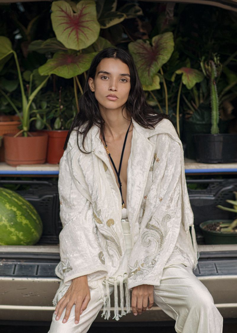 A young adult female model poses while seated with an embroidered coat, plants, and a watermelon in the background, medium shot, indoor.
