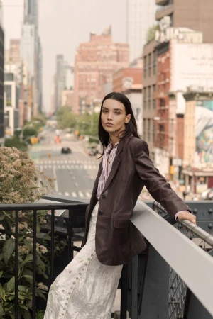 A stylish woman poses in a brown blazer and patterned skirt on an elevated walkway in a city, with tall buildings in the background.