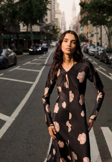 Elegant woman in black long-sleeved dress with seashell pattern stands on a city street with tall buildings and a crosswalk.