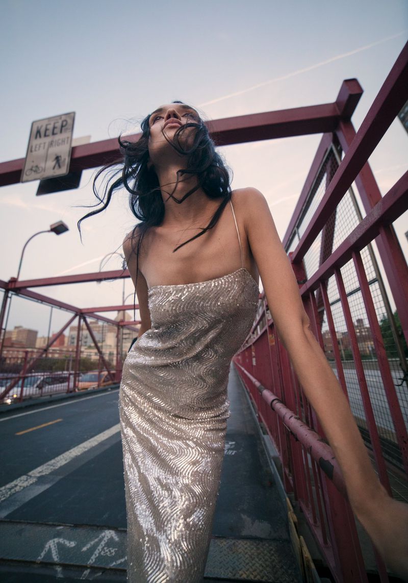 Brunette woman in a sequin dress poses on a red bridge with city buildings in the background; fashion editorial on an overcast day.