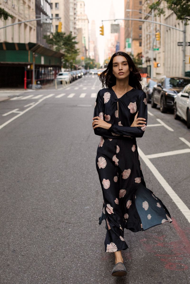 Woman in patterned dress walks down city street with tall buildings and cars during daytime, showcasing fashion and urban style.