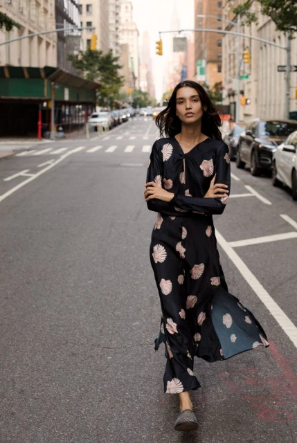 Woman in patterned dress walks down city street with tall buildings and cars during daytime, showcasing fashion and urban style.