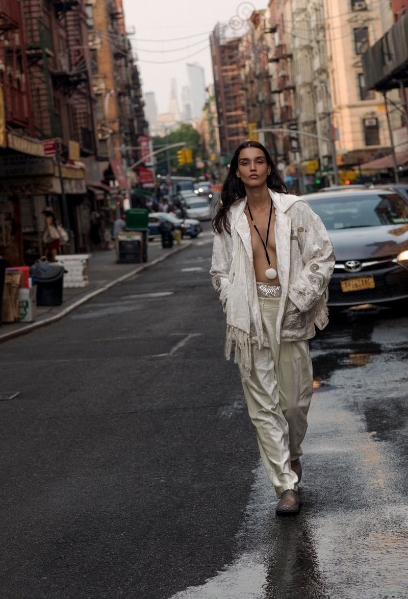 Adult walks down a wet city street lined with buildings and parked cars, wearing a patterned jacket and light-colored pants.