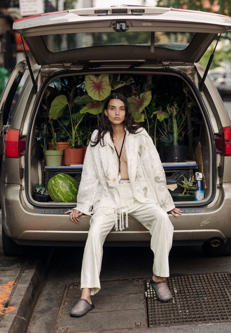 A woman sits on the open trunk of a car filled with potted plants, wearing an off-white suit with textured jacket and dark footwear in daylight.