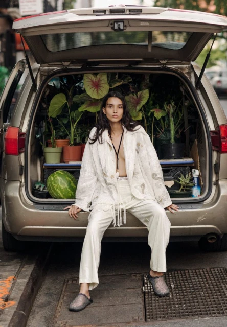 A woman sits on the open trunk of a car filled with potted plants, wearing an off-white suit with textured jacket and dark footwear in daylight.