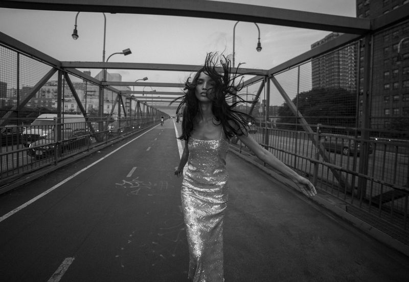 A slender adult female with long dark hair wearing a sparkling gown, walks on an outdoor overpass with buildings in the background.