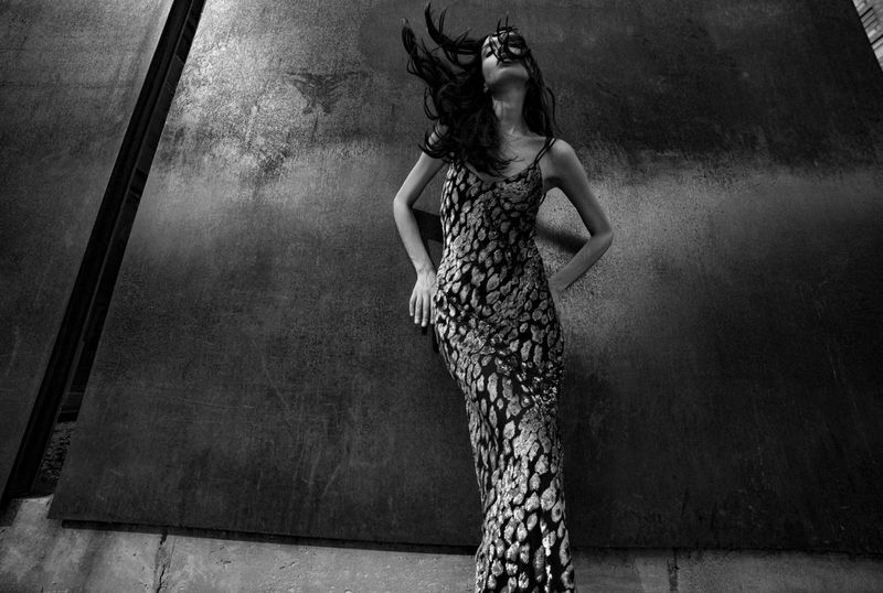 Adult woman poses in a dress against a dark background, hair blowing, fashion photography in black and white, studio shot.