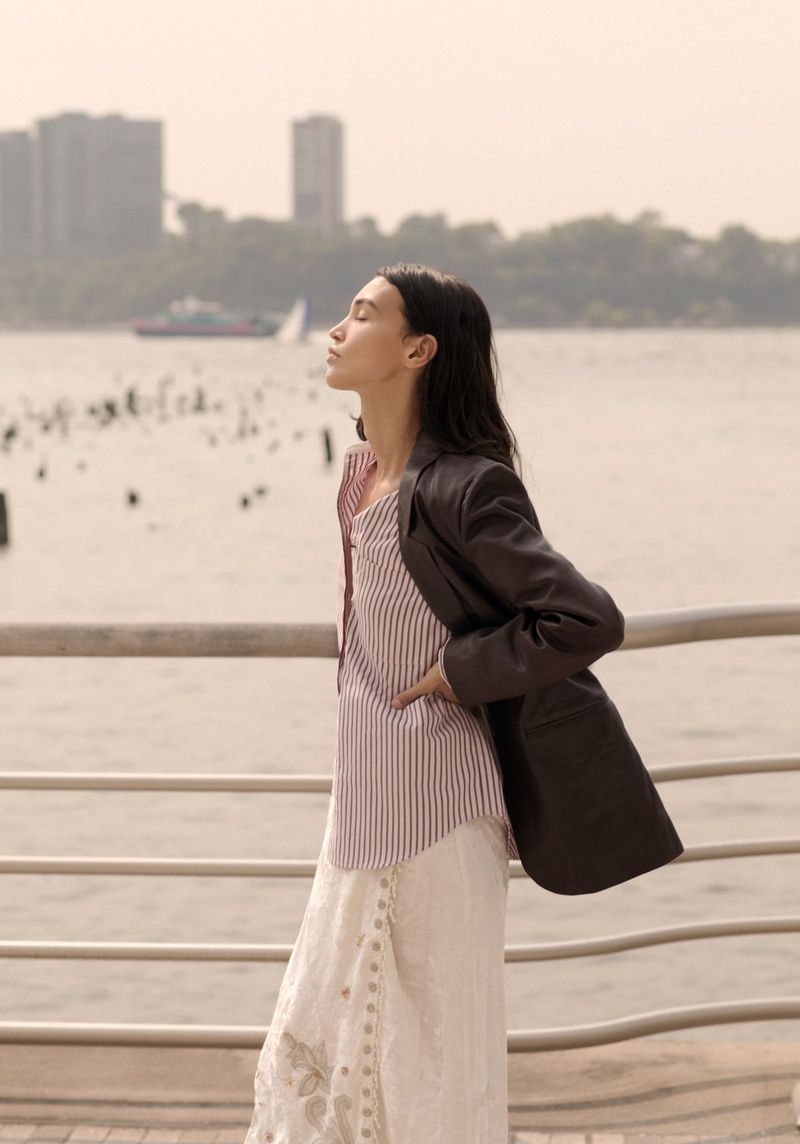 Woman in blazer and striped top stands by waterfront with sunlight on face. Urban background with boat on water.