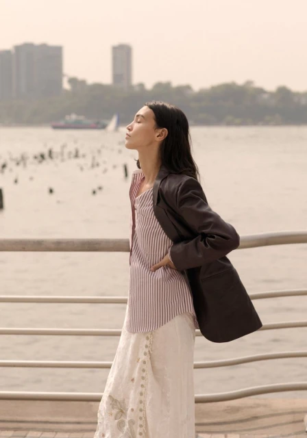 Woman in blazer and striped top stands by waterfront with sunlight on face. Urban background with boat on water.
