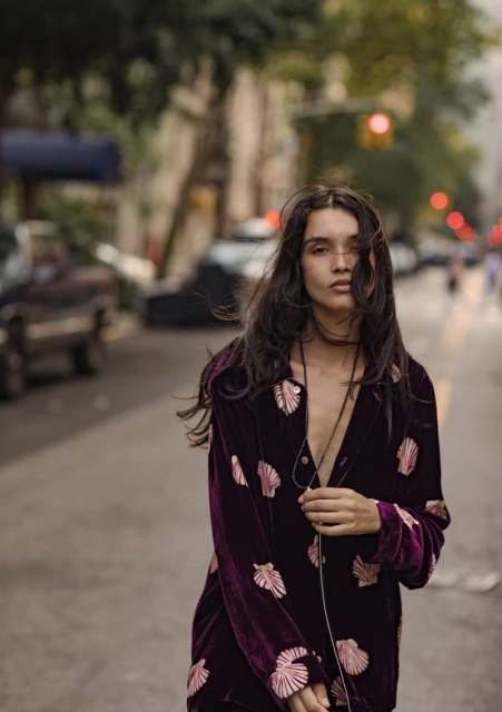 Woman in a purple velvet seashell-print jacket poses on a city street with blurred cars and buildings in the background during daytime.