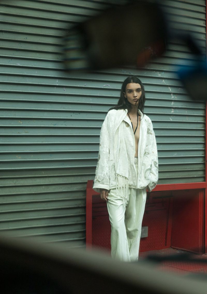 A woman poses in white jacket and pants against a grey horizontally-lined wall with red structural accents in an outdoor setting.