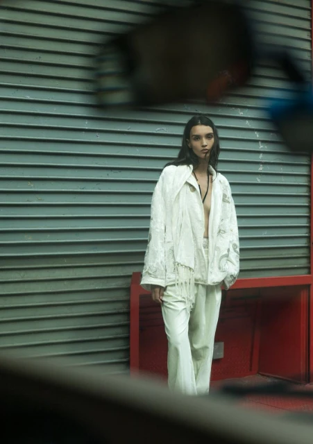 A woman poses in white jacket and pants against a grey horizontally-lined wall with red structural accents in an outdoor setting.