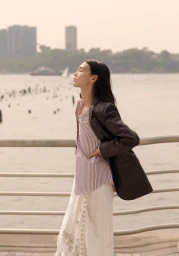 Model standing outdoors by a waterfront railing wearing a dark blazer, striped top, and patterned white skirt.