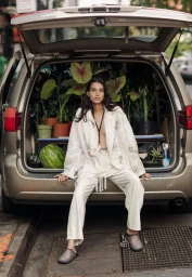 Model seated in the open trunk of a car filled with potted plants and a watermelon, wearing a white patterned jacket and trousers.