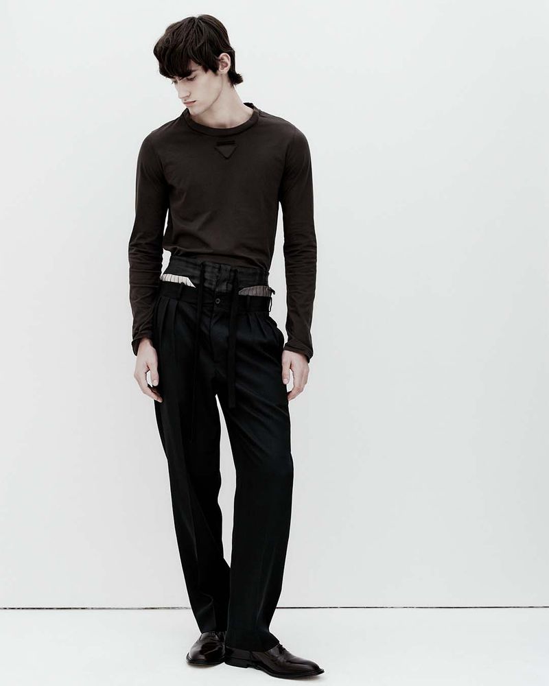 Male fashion model poses in a brown long-sleeved shirt and dark pleated trousers against a plain white backdrop in a studio setting.