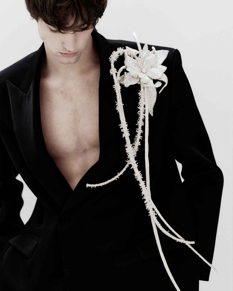 Man in a black suit jacket with a white floral brooch, showcasing fashion and style against a plain white background in a studio setting.