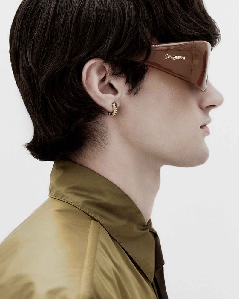Profile portrait of a male model wearing dark wrap-around sunglasses and an olive green collared shirt against a white background.