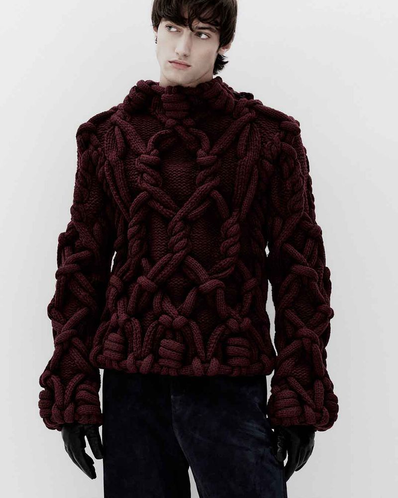 Portrait of a young adult man wearing a burgundy, rope-knit wool sweater and black gloves against a plain, light grey backdrop.