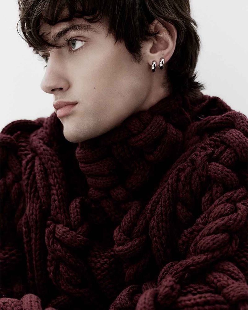 Profile shot of a young adult male with dark hair wearing a chunky knitted maroon sweater, looking to the side against a plain white backdrop.
