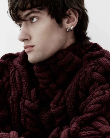 Close-up portrait of a young male model wearing a thick textured burgundy turtleneck sweater and silver hoop earrings against a white background