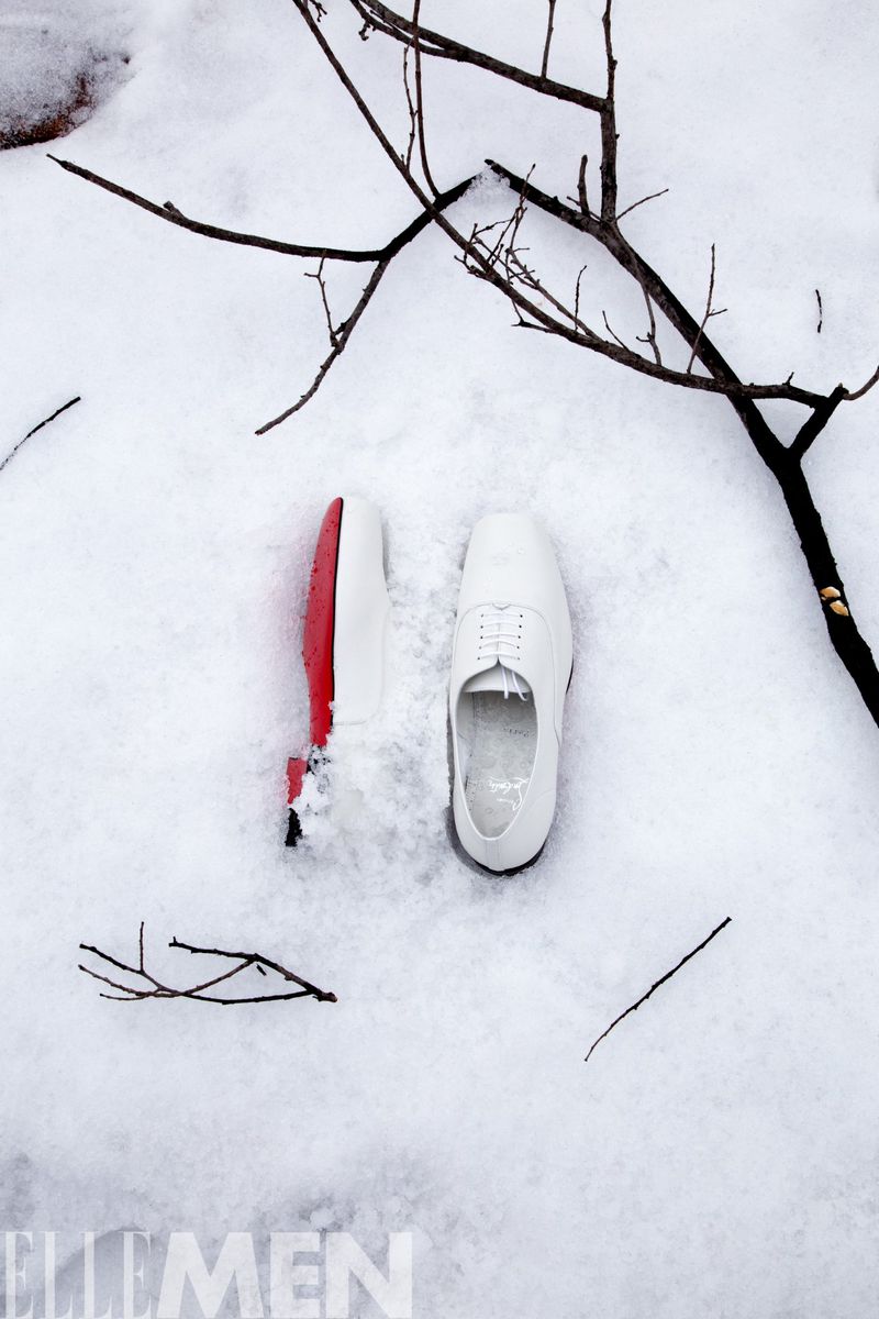 Two white dress shoes, one with a red sole, are displayed on fresh snow with bare tree branches in the background. Winter fashion.