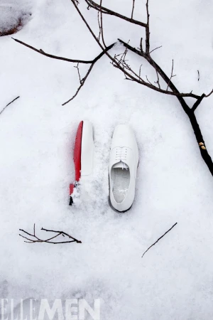 Two white dress shoes, one with a red sole, are displayed on fresh snow with bare tree branches in the background. Winter fashion.