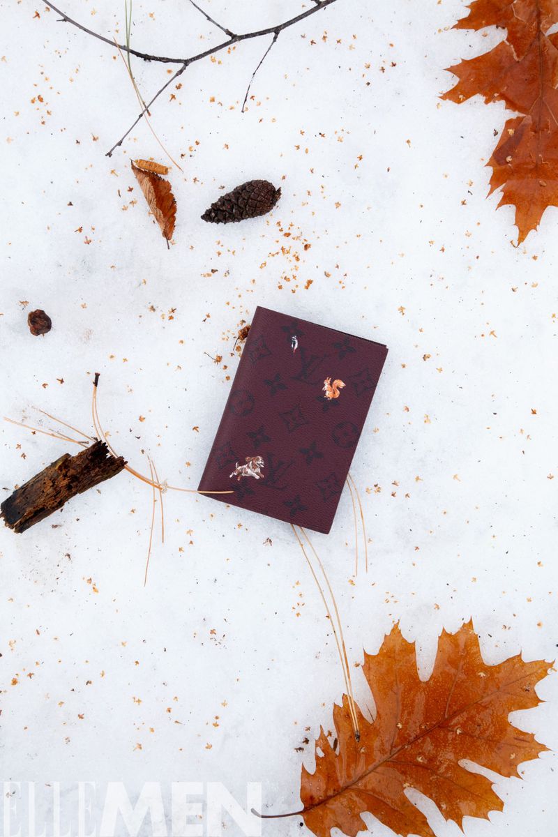 Burgundy notebook on white snow with scattered brown leaves and pine needles, creating a serene and wintry outdoor scene.