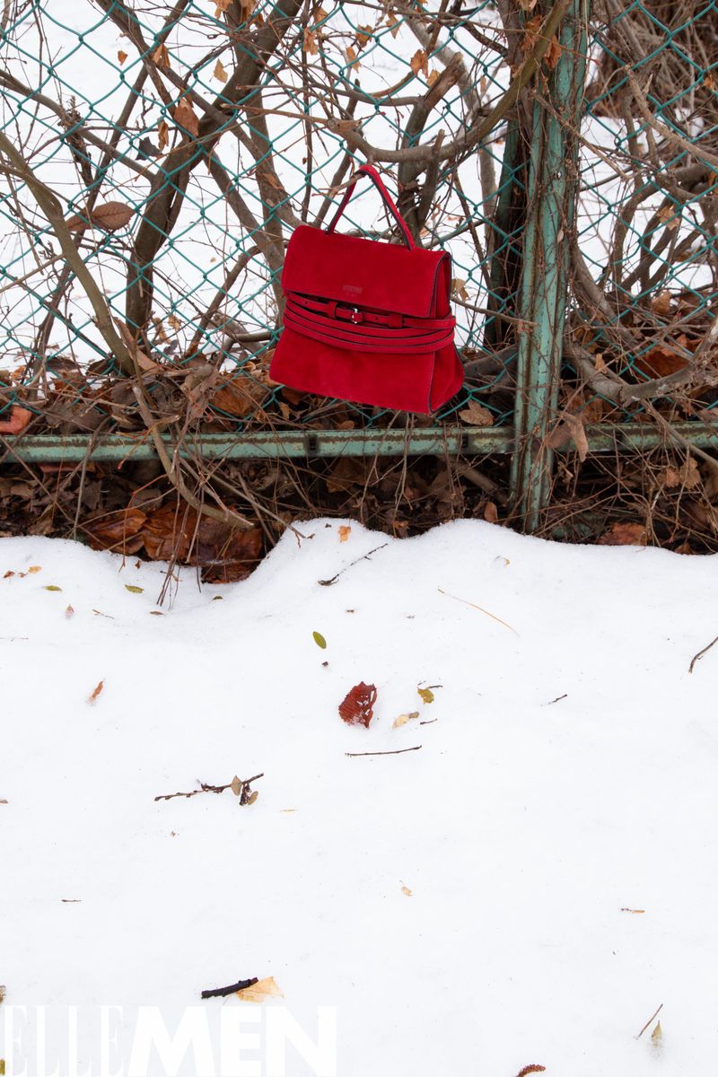 Red suede handbag hanging on a chain-link fence covered in snow, surrounded by bare branches and leaves in an outdoor setting.