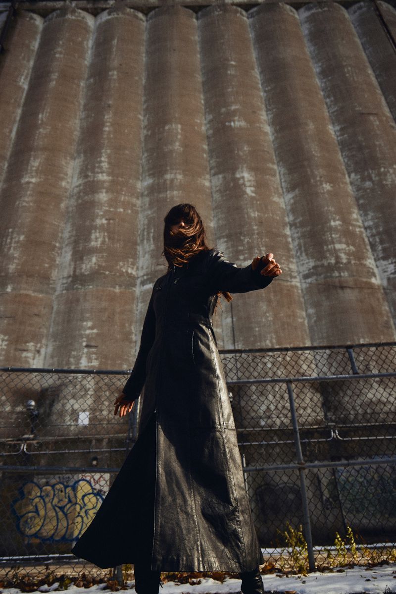 A woman in a long black coat stands in front of a tall concrete silo with graffiti near the bottom of the structure.