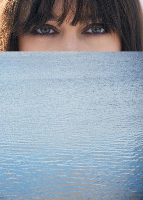 Close-up of a woman's face with blue eyes and dark hair is partially obscured by a blue ocean water background in an eye-level portrait.