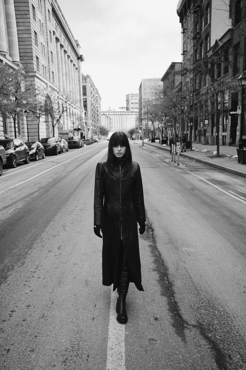 A woman in a black coat walks down an urban street in black and white, framed by tall buildings and cars, creating a striking fashion image.