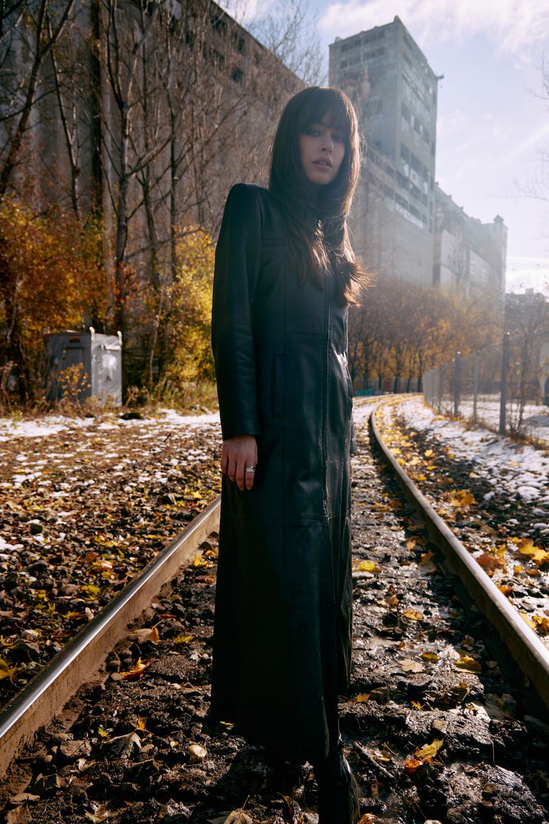 A woman in a long black coat stands on railroad tracks surrounded by fallen leaves and an industrial building on a sunny day.