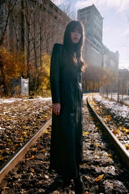 A woman in a long black coat stands on railroad tracks surrounded by fallen leaves and an industrial building on a sunny day.