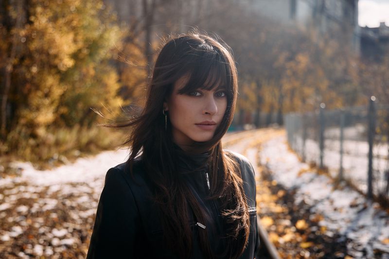 Portrait of a brunette woman with bangs in a black jacket, posing outdoors in a park setting with autumn foliage in the background.