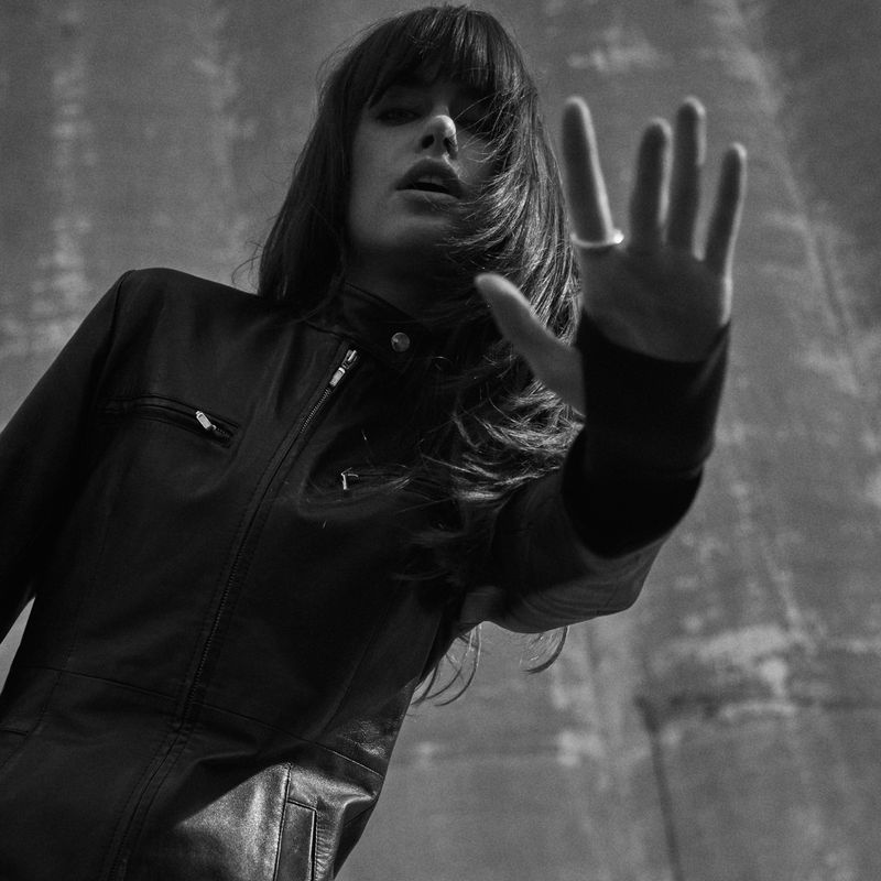 Monochrome portrait of a young woman in a black leather jacket raising her hand in front of a textured grey concrete wall.