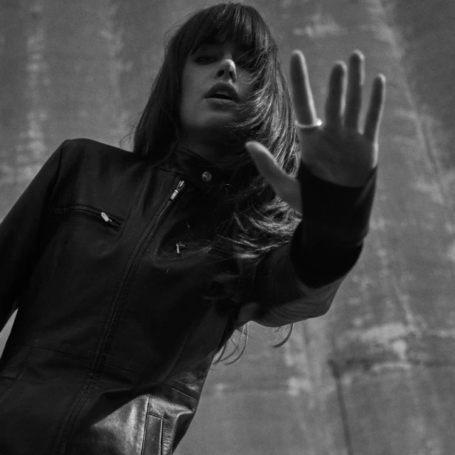 Monochrome portrait of a young woman in a black leather jacket raising her hand in front of a textured grey concrete wall.