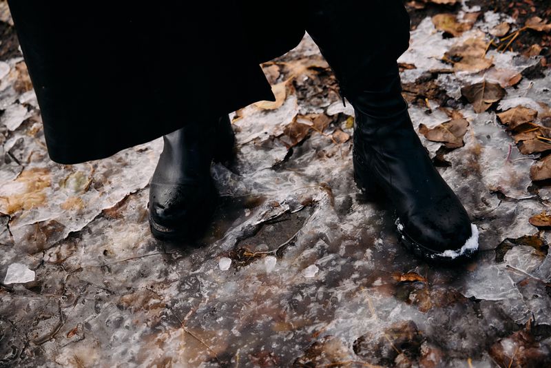 Close-up of black leather boots standing on a frozen icy puddle with scattered fallen leaves.