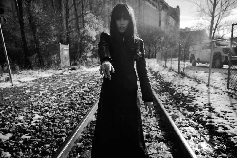Black and white shot of woman in long coat walking on snow-covered train tracks in an outdoor setting, with bare trees and a building.