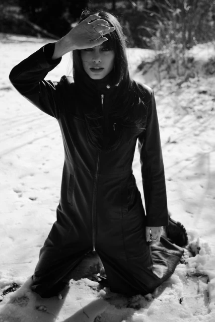 Woman in a black jumpsuit kneels in the snow, shielding her eyes from the sun; outdoor fashion portrait with a shallow depth of field.
