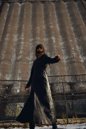 Model wearing a long black leather coat outdoors in front of tall industrial silos and a chain-link fence.