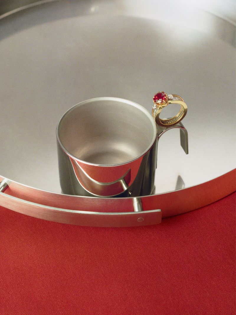 Gold ring with red gemstone on silver cup handle, silver tray, and red fabric background in a still life product shot.