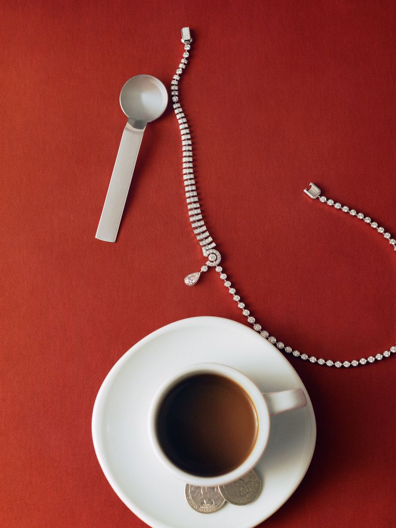 Overhead shot of coffee, saucer, silver spoon, two coins, and diamond necklace on a vibrant red surface. Jewelry and drink still life.